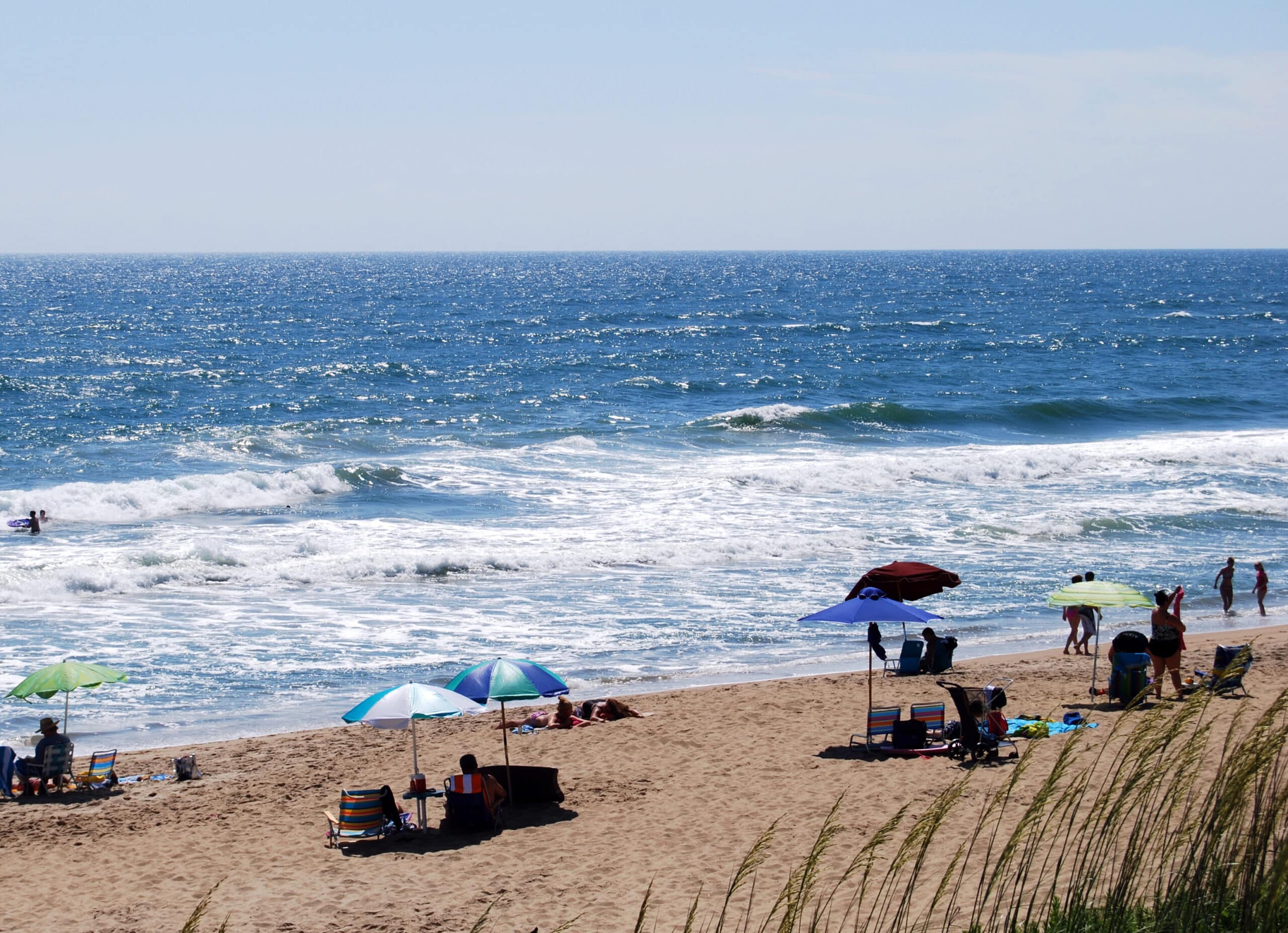 Rip current visible on an Outer Banks beach as a dark gap in the breaking waves, with beachgoers under colorful umbrellas and sea oats in the foreground.
