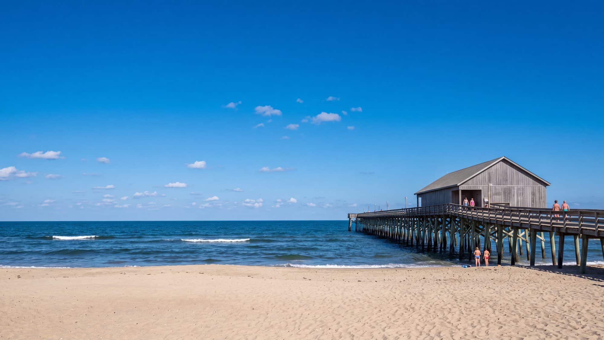 A wooden fishing pier with a pier house seen from the beach on a bright day with a few swimmers — Outer Banks, NC