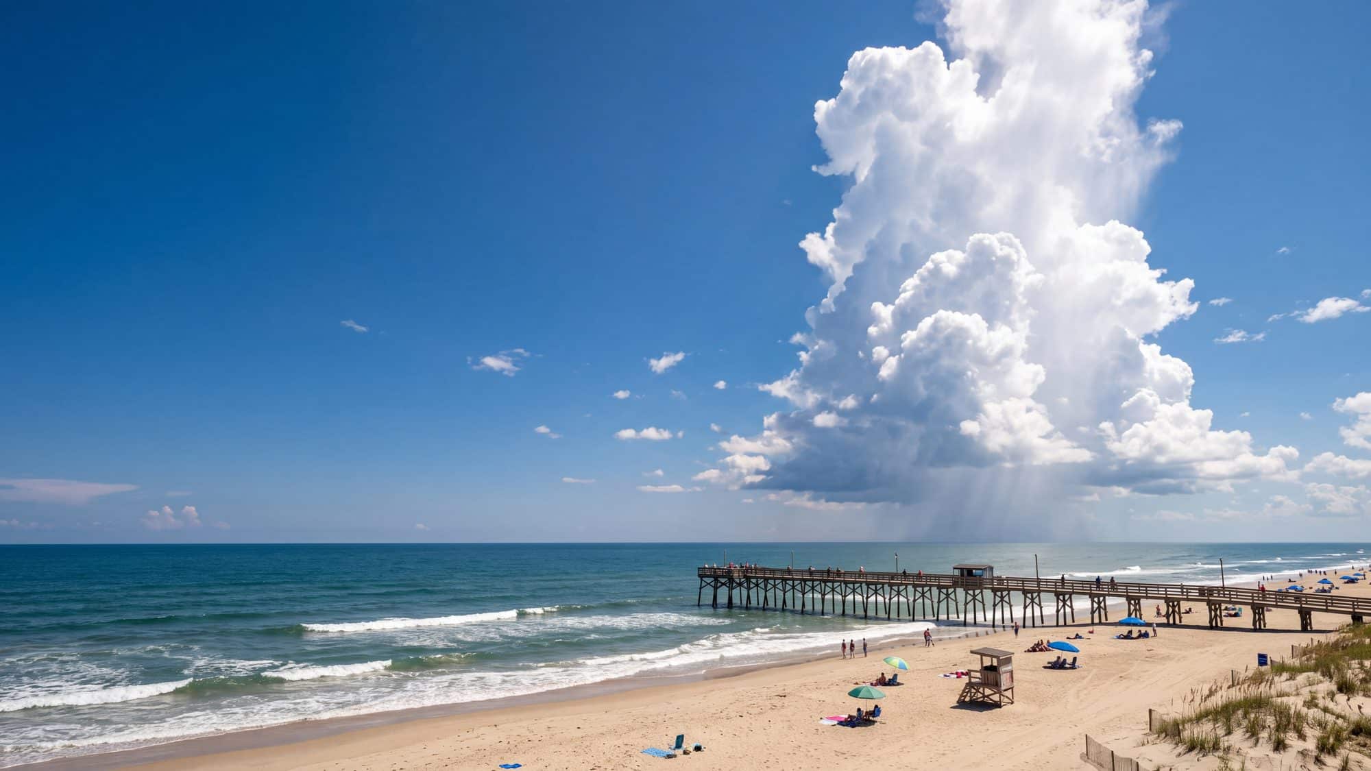 A towering afternoon cumulus cloud over a wide beach with a fishing pier and bright sun breaking through — Outer Banks, NC