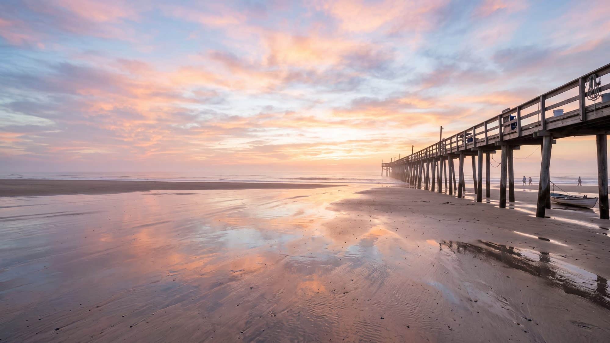A wide flat beach at low tide with the wet sand mirroring a colourful sky, a wooden fishing pier far down the beach — Outer Banks, NC