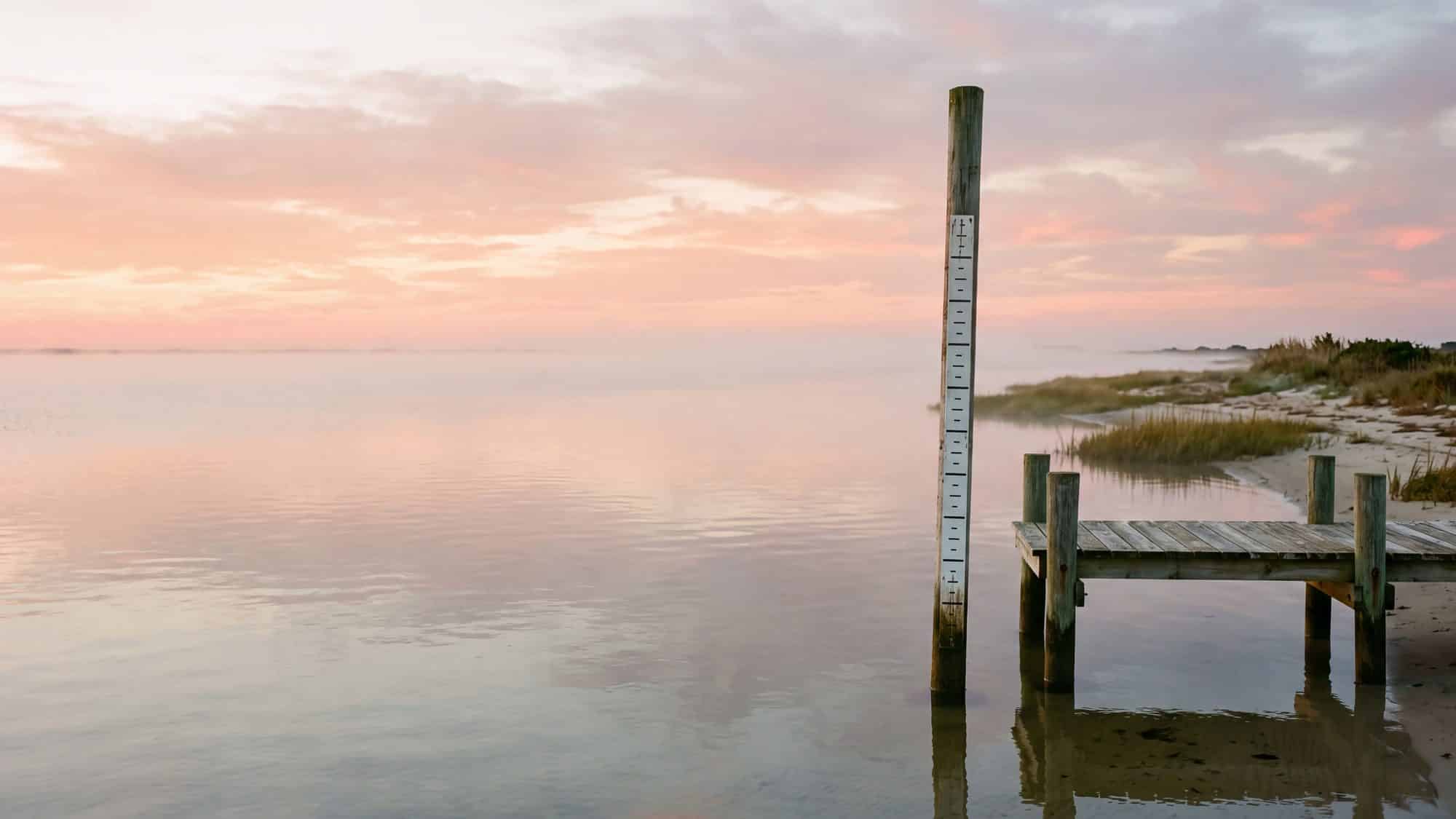 A tall graduated tide staff pole standing in calm water beside a wooden dock — Outer Banks, NC