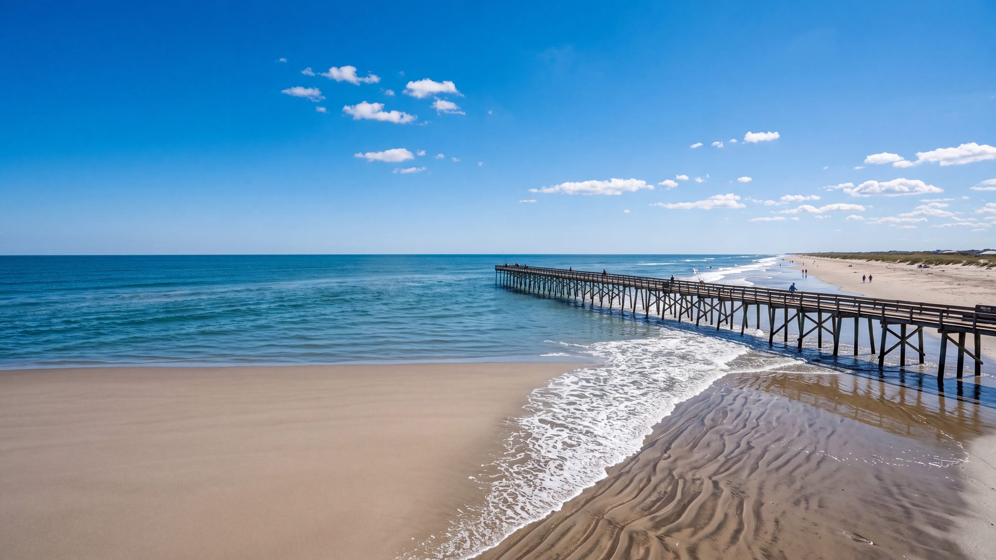 A wide beach at mid tide with waves washing over ridged sand and a pier far off — Outer Banks, NC